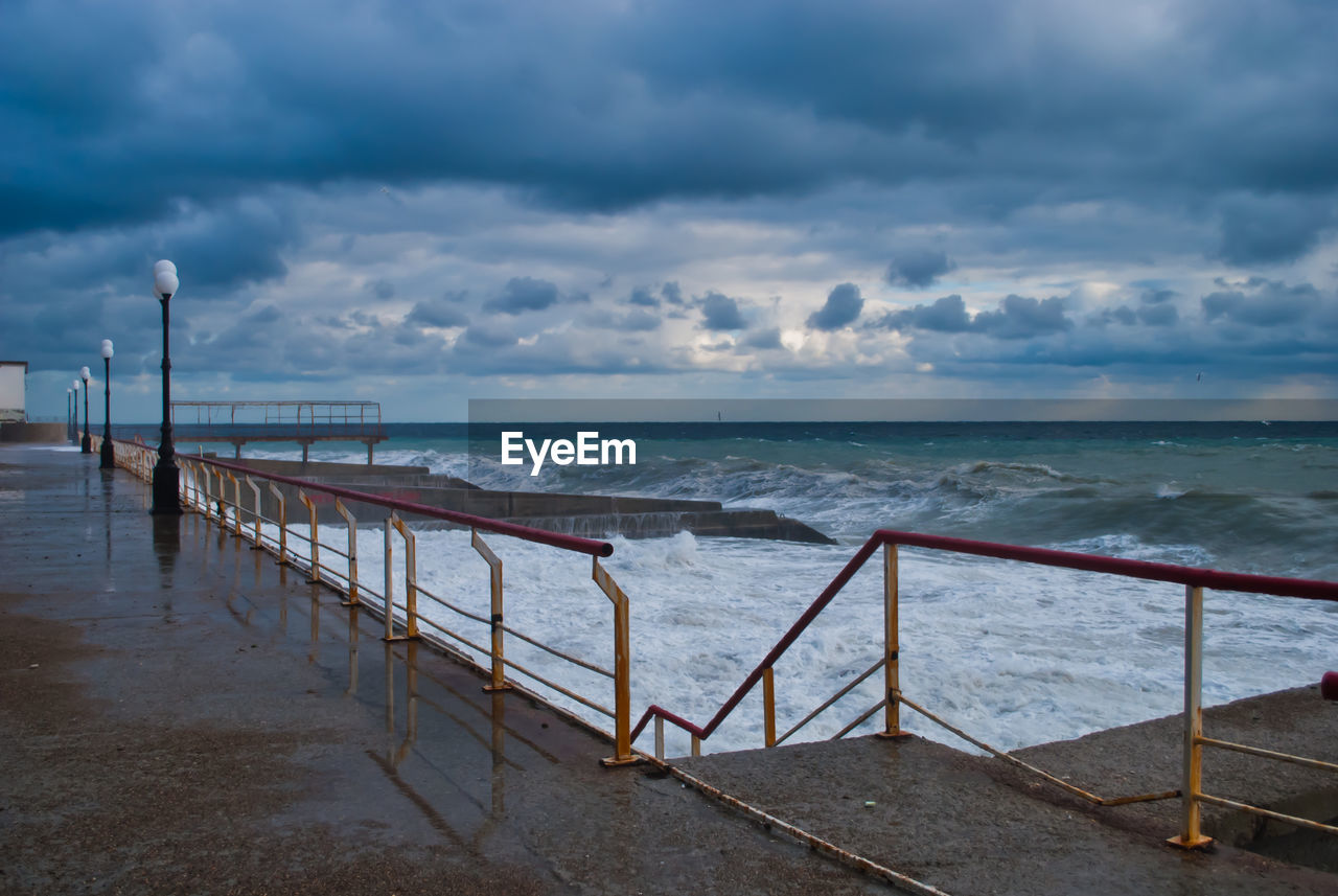 Street lights on pier with waves splashing in sea against cloudy sky