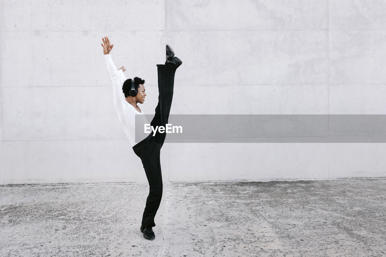 Flexible male dancer stretching leg while practicing urban dance against white wall