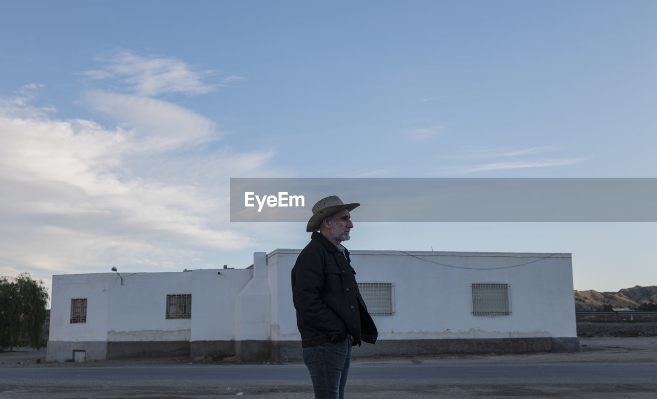 Adult man in cowboy hat standing in front of white building against sky