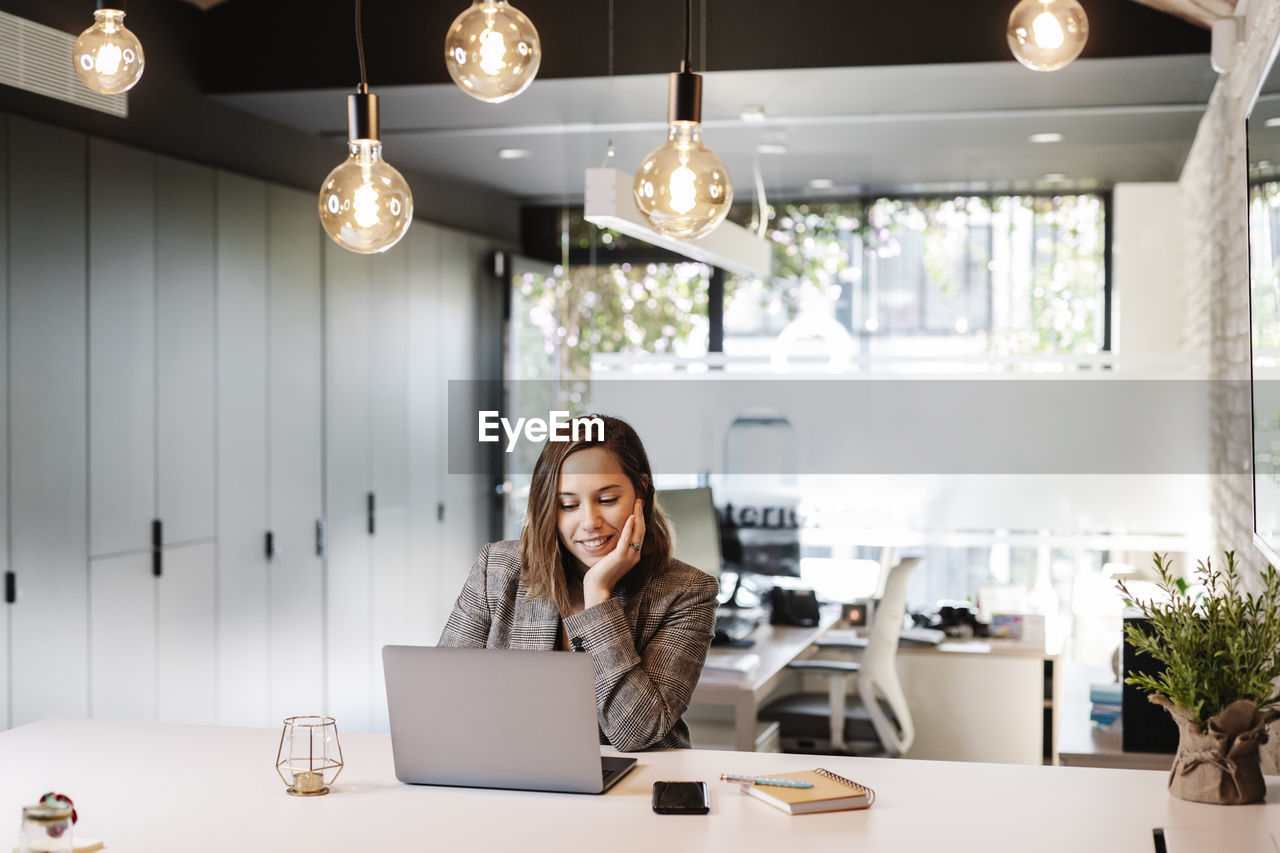 Smiling businesswoman with hand on chin using laptop at desk in office