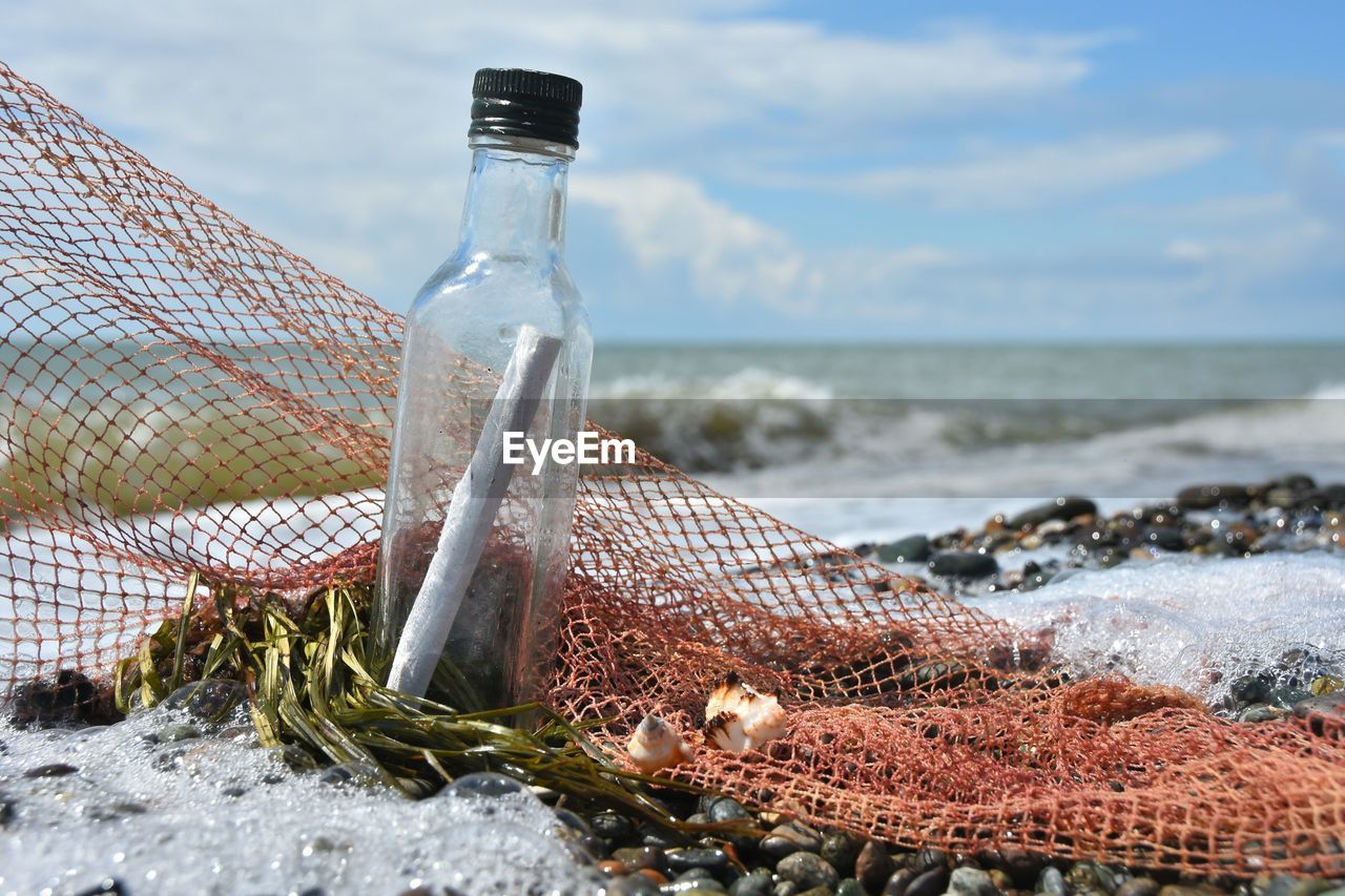Close-up of message in bottle by fishing net at beach against sky
