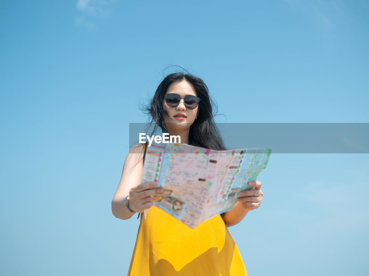 Smiling young woman holding map while standing against blue sky
