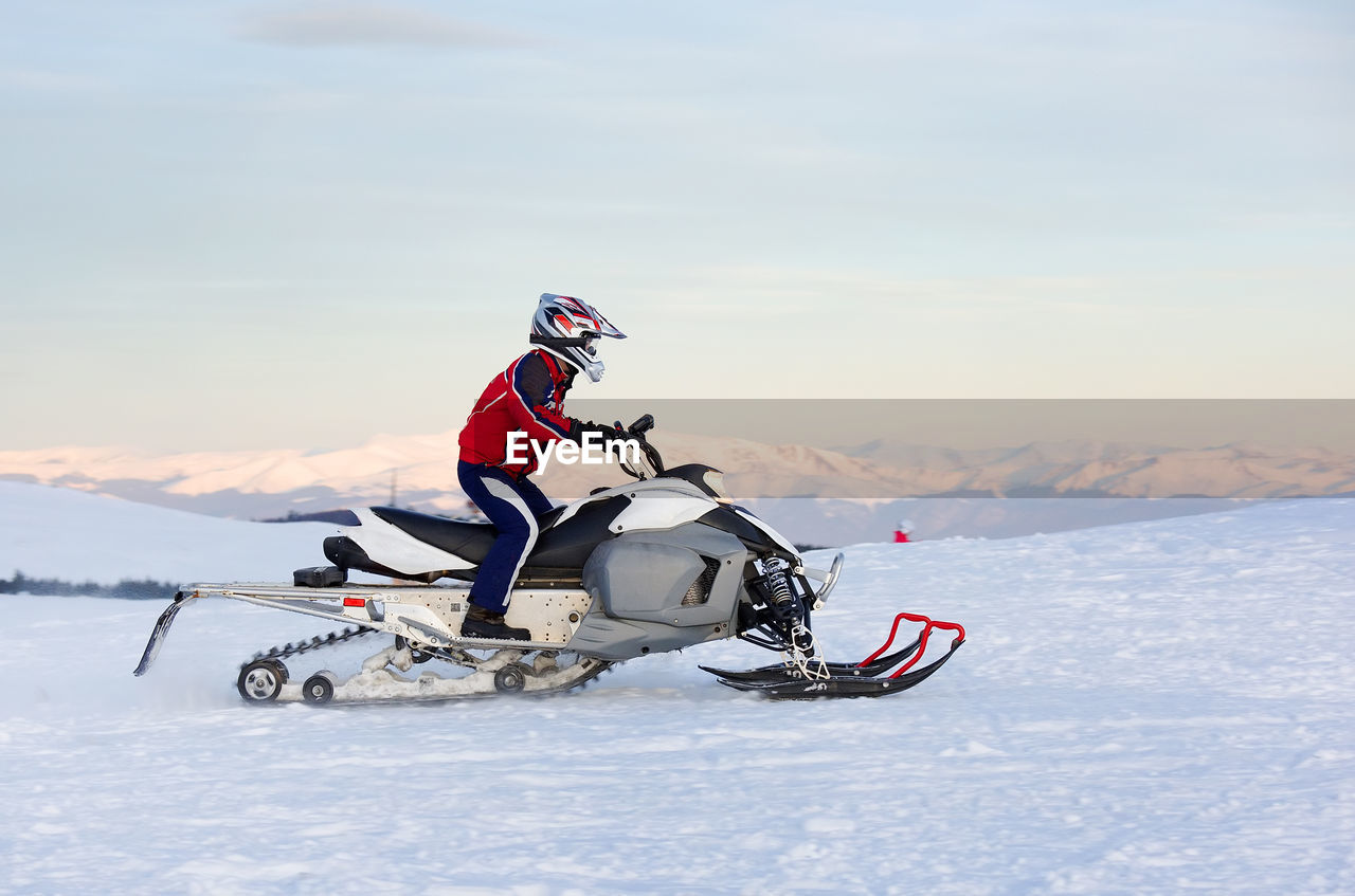 Side view of man riding snowmobile on snow covered landscape against sky