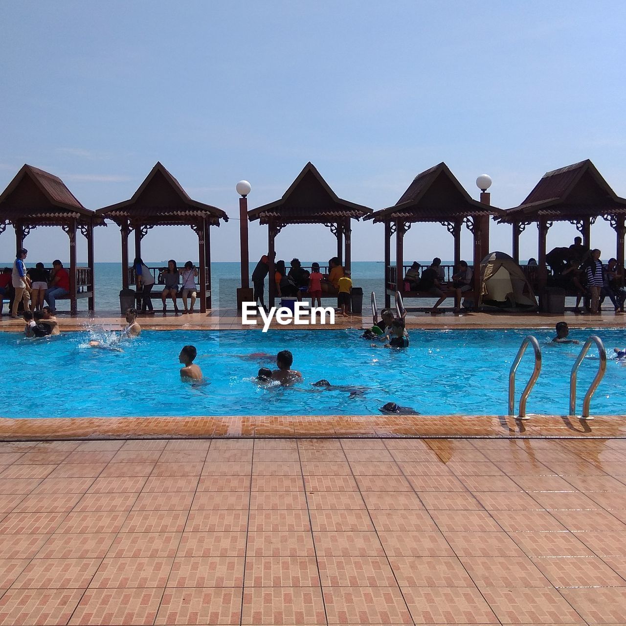 GROUP OF PEOPLE AT SWIMMING POOL AGAINST CLEAR BLUE SKY