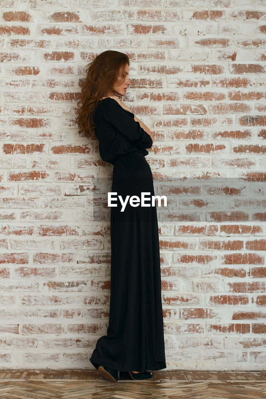 Full length of beautiful woman in black dress standing against brick wall