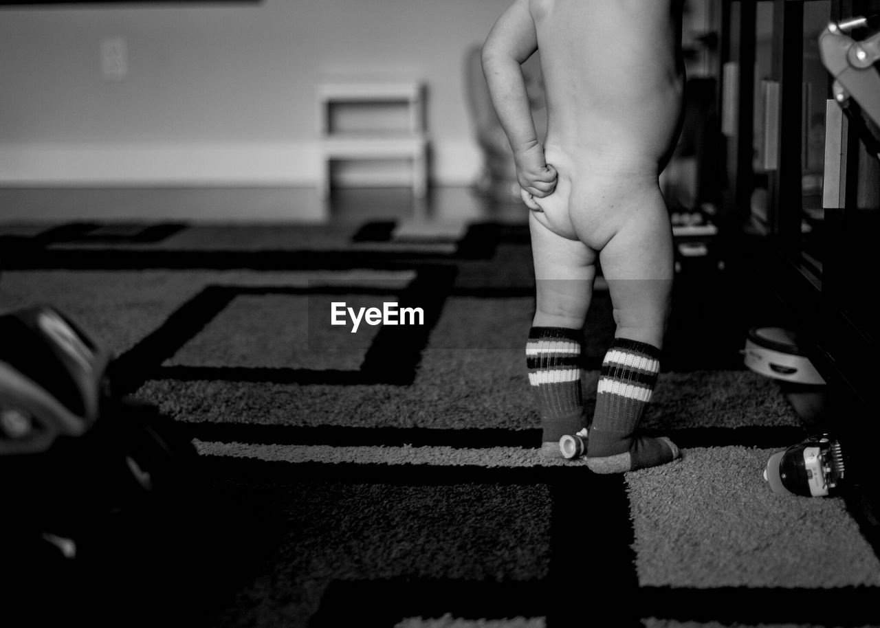 Low section of naked baby boy scratching buttocks while standing on carpet at home