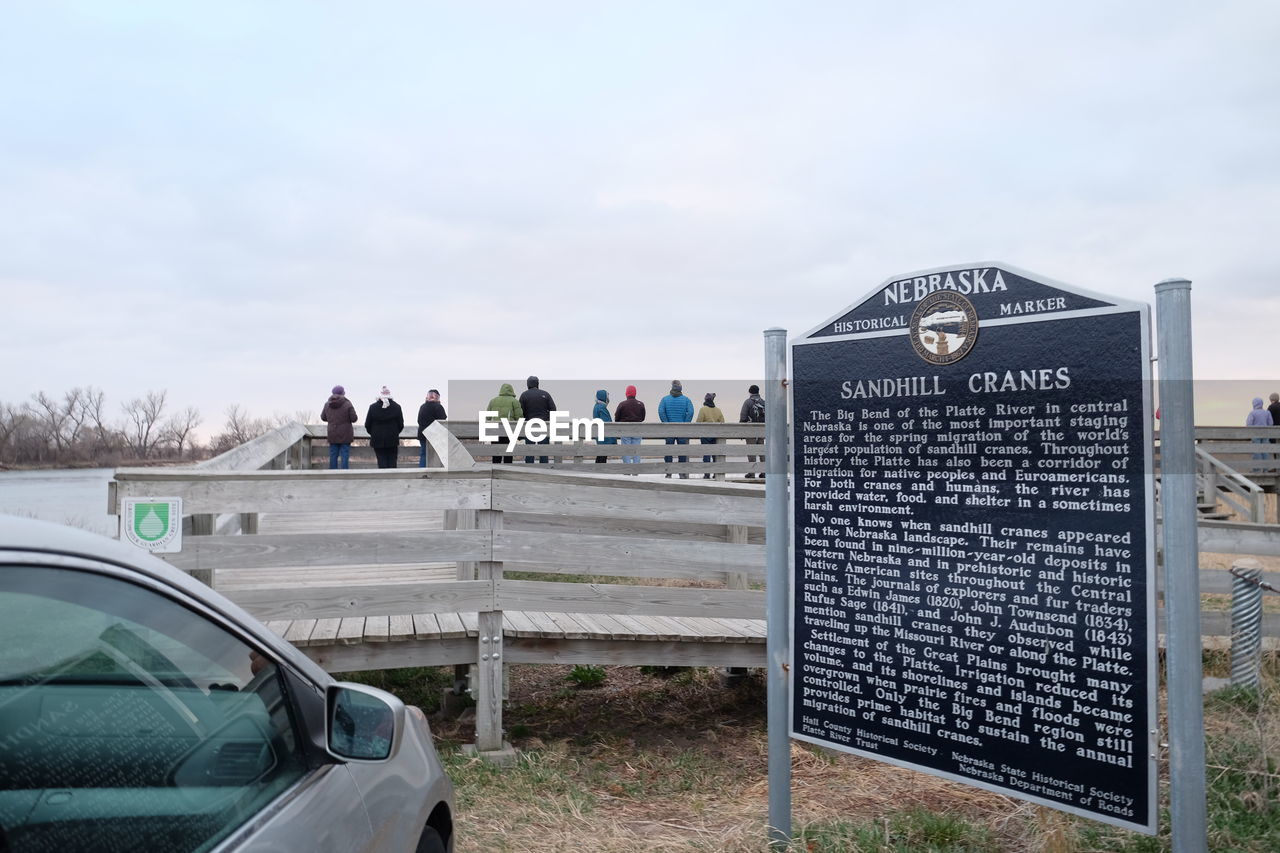 Car parked by historical marker with people on pier against sky