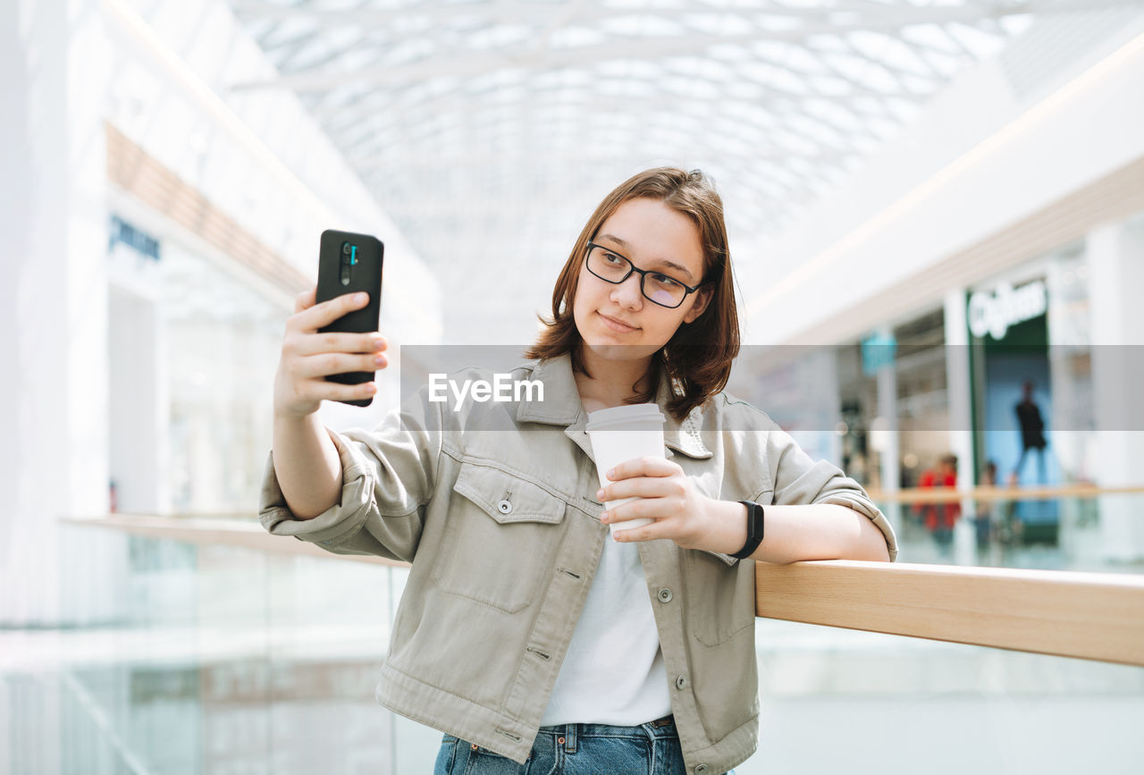 Young teenager girl student in glasses using mobile phone taking selfie at shopping mall