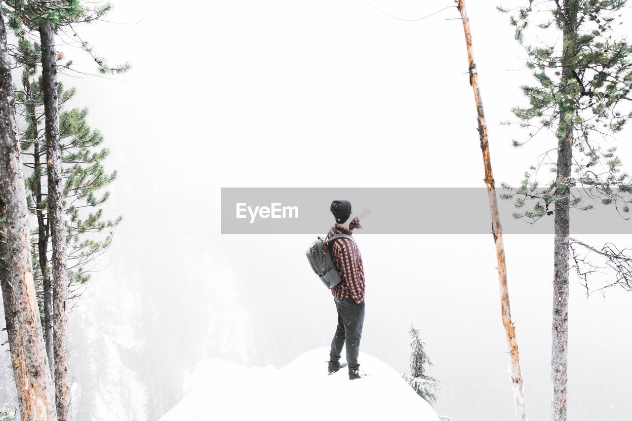 Side view of man with backpack standing on snow in forest during foggy weather