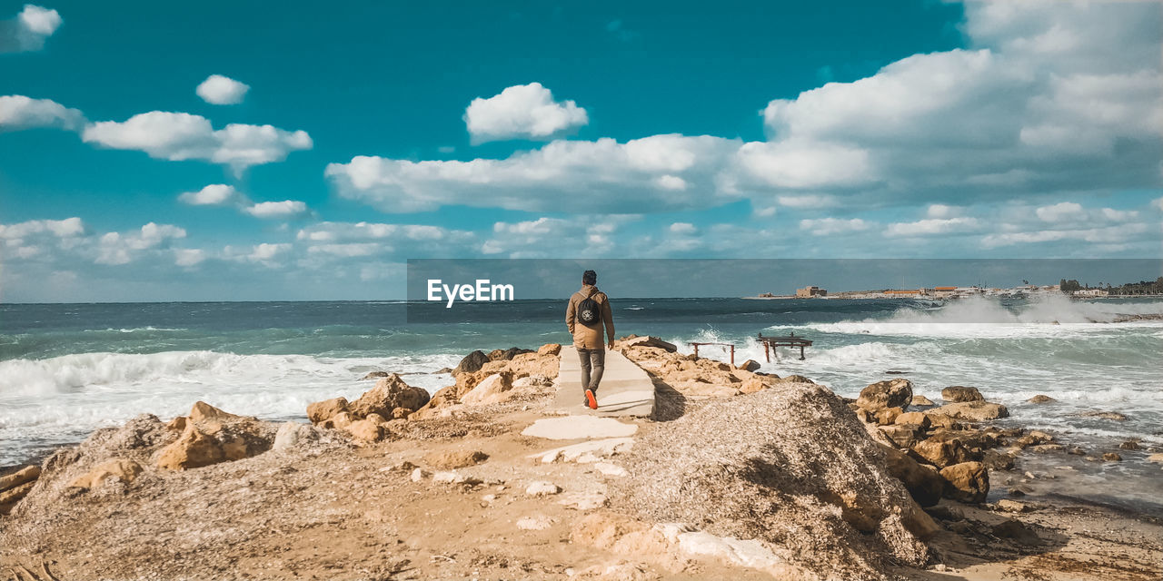 MAN STANDING ON ROCKS AT BEACH AGAINST SKY