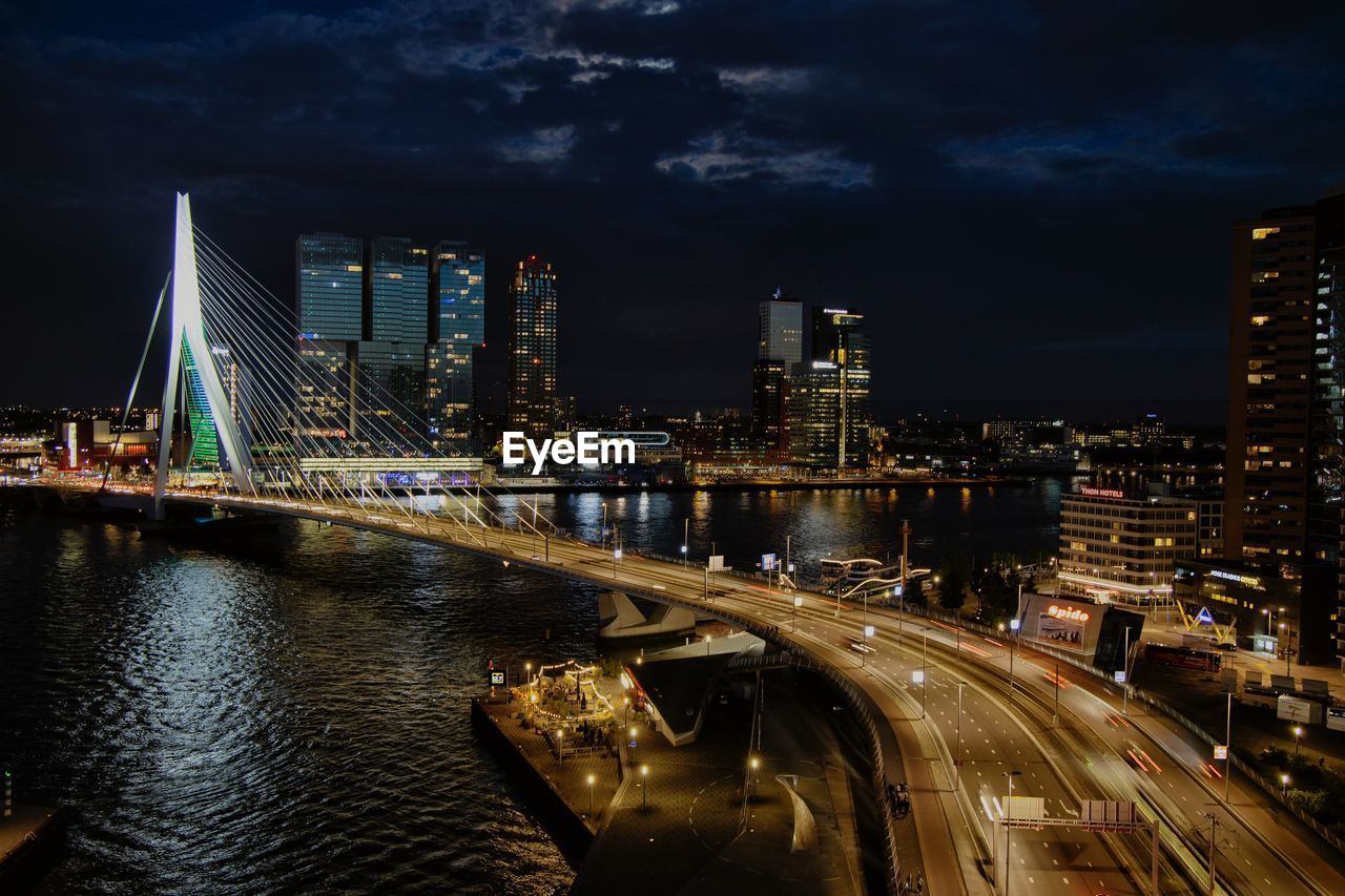 Illuminated bridge over river by buildings against sky at night