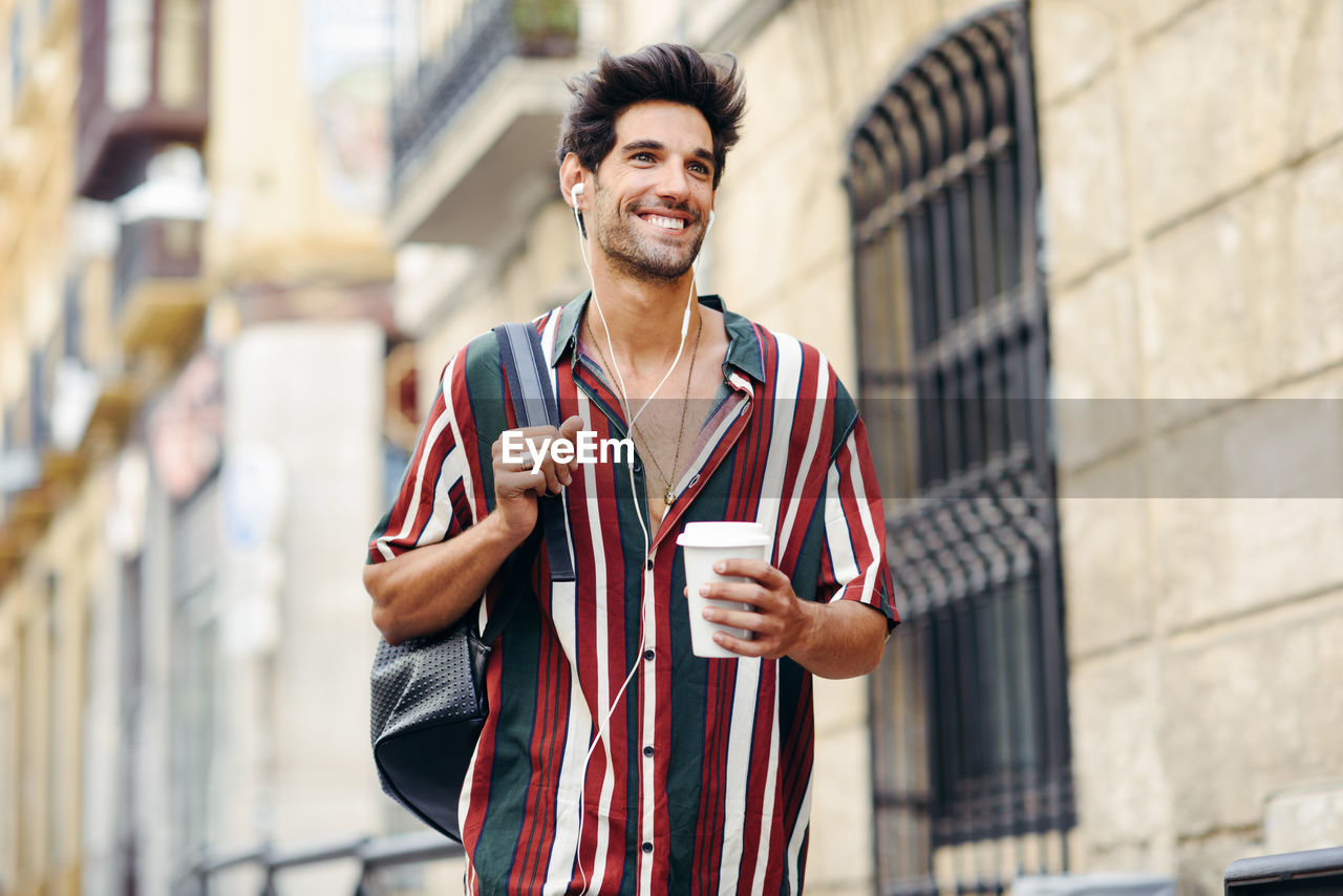 Smiling man holding disposable cup while standing against building in city