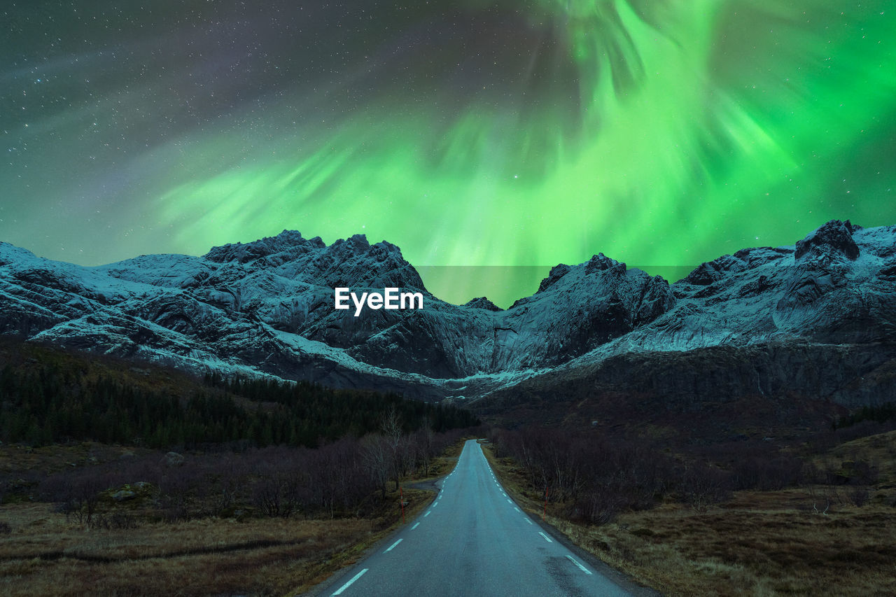 Scenic view of automobile driving on empty mountainous road in winter under night sky glowing with bright green aurora borealis in norway