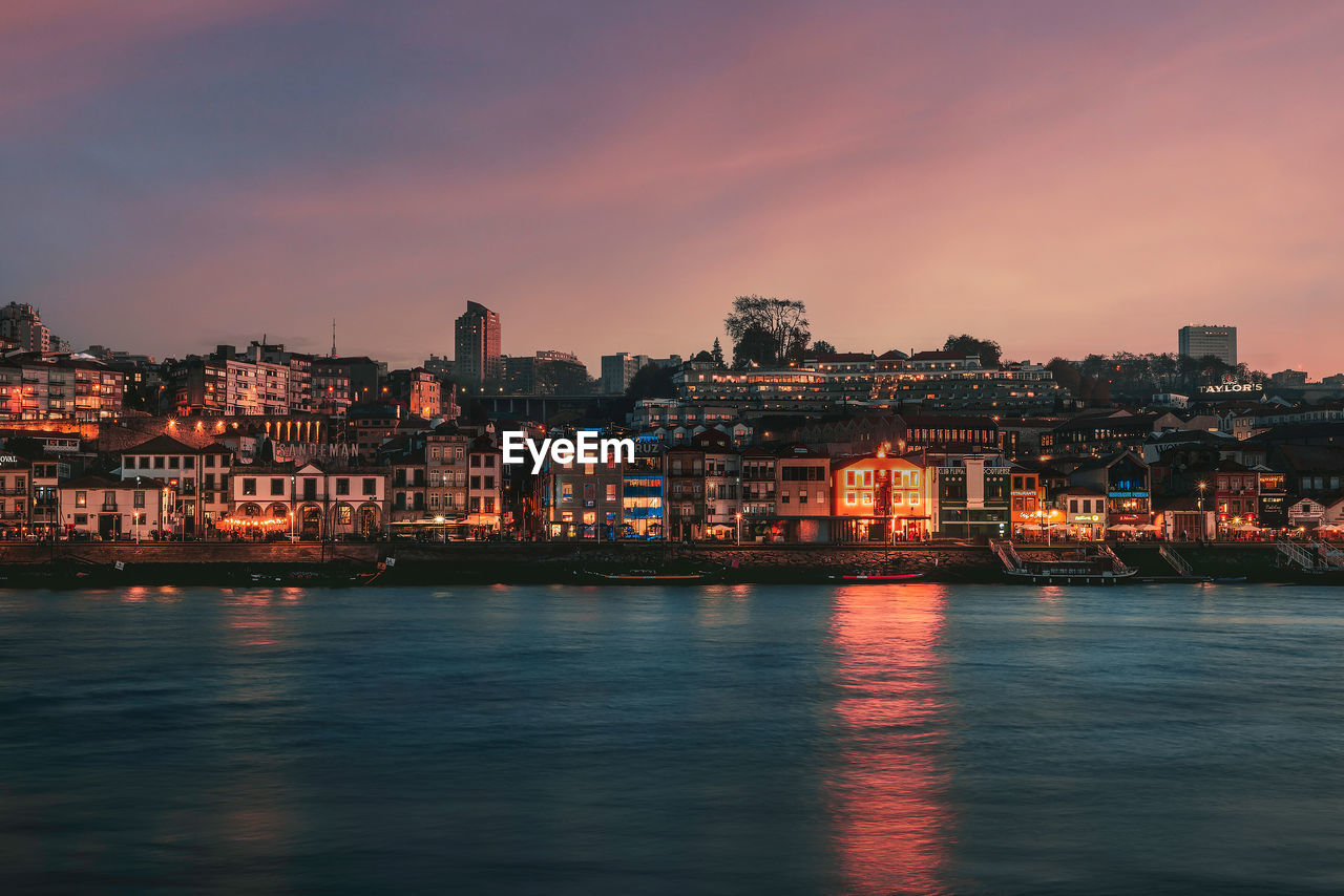 Illuminated buildings by river against sky at sunset