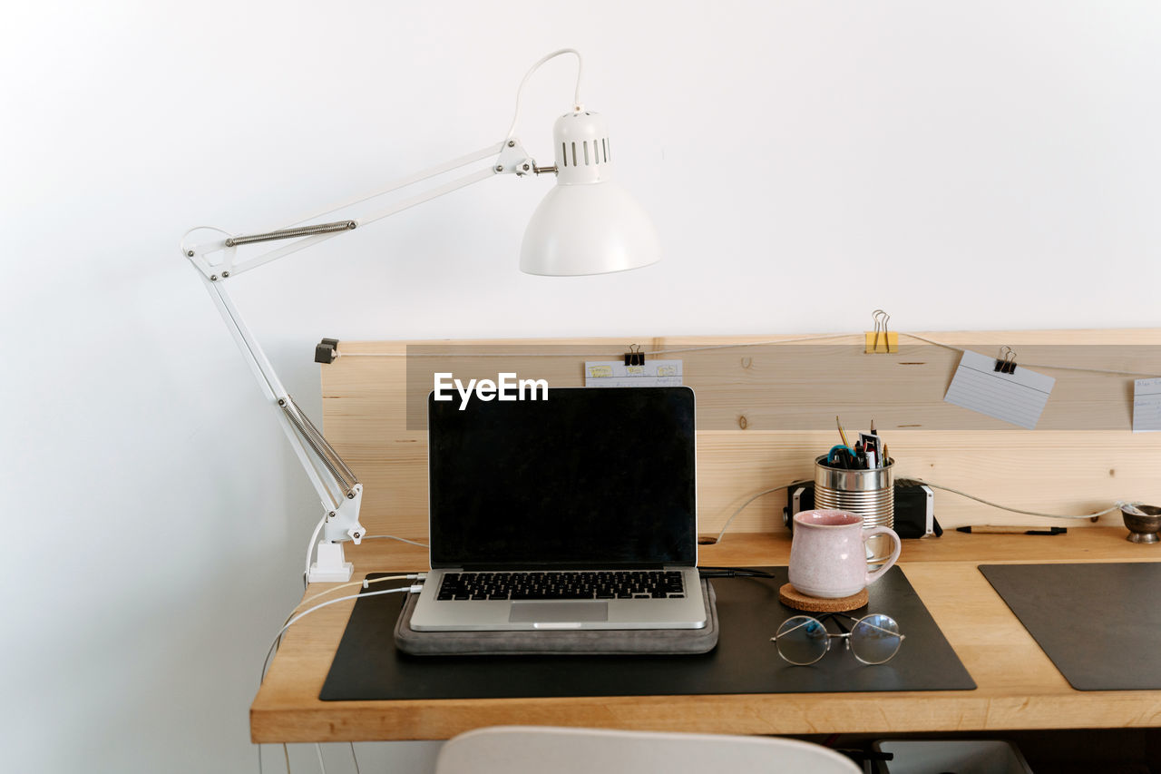 From above composition of modern portable netbook with black screen placed on wooden desk near cup and eyeglasses in light office