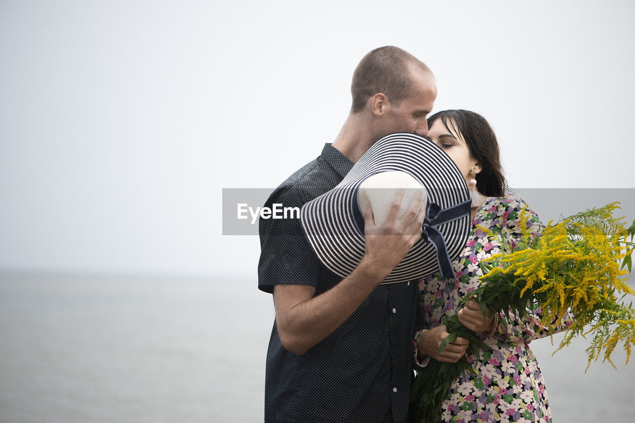 Young romantic couple kisses and hugging, hiding their faces behind their summer hats