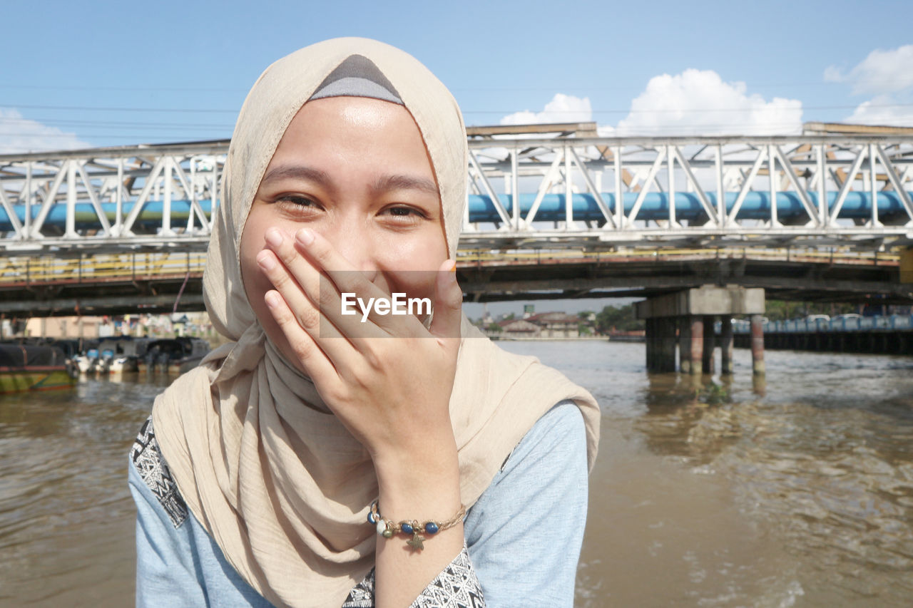 Portrait of happy young woman standing by river against sky