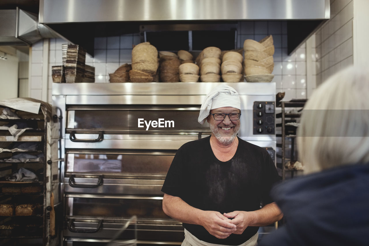 Smiling senior male baker standing against cooling rack at bakery