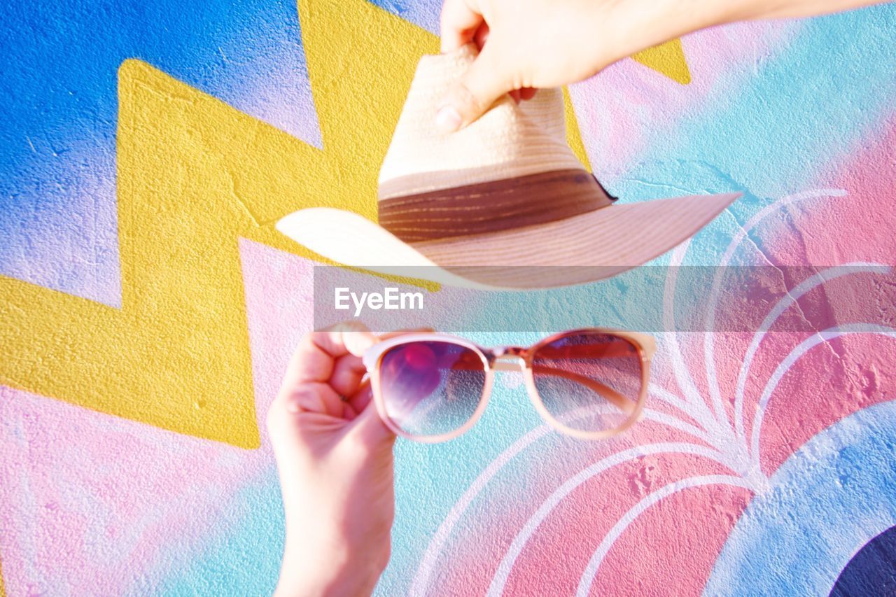 Cropped hands holding hat and sunglasses against patterned wall