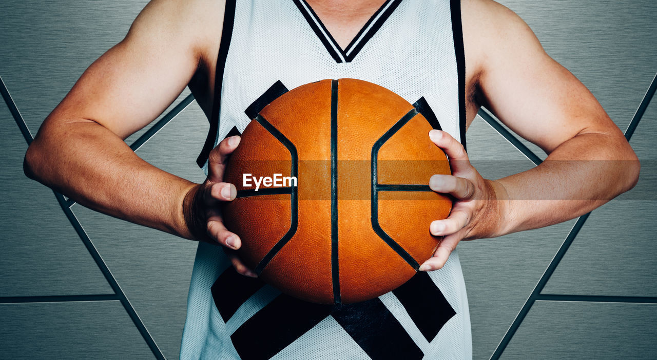 Midsection of sportsman holding basketball while standing against wall