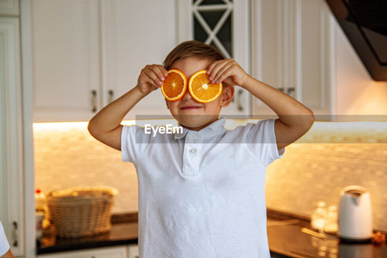 A funny boy holding fresh orange slice on eyes in kitchen