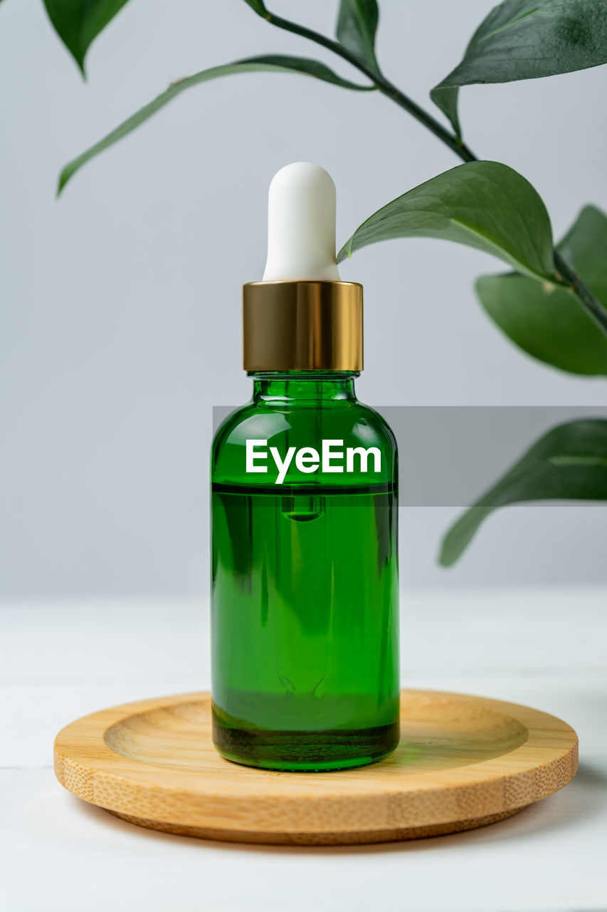Green cosmetic bottle with pipette on wooden podium, product packaging with natural ruscus branch