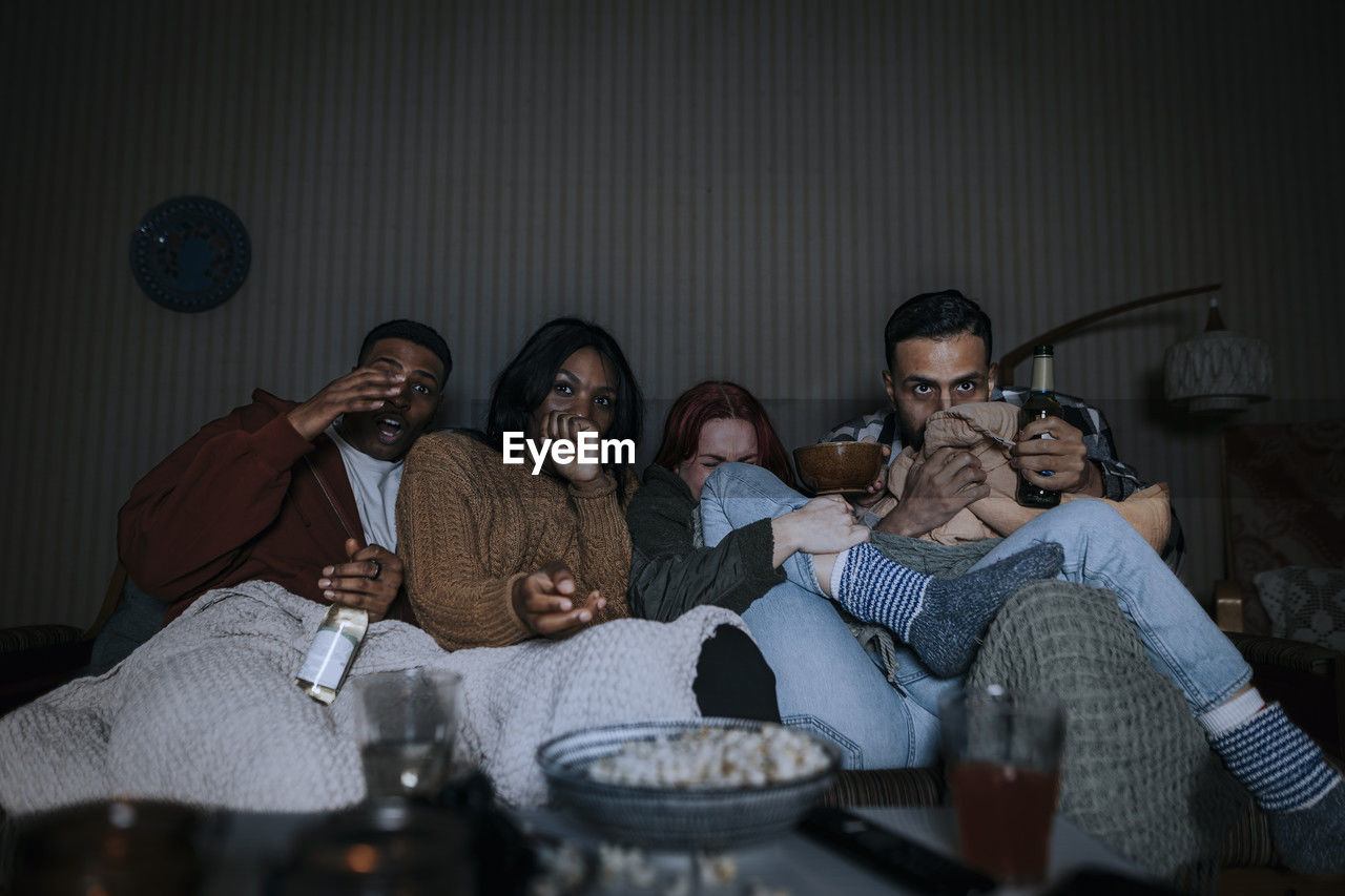 Multiracial friends watching scary movie together while sitting on sofa in cabin