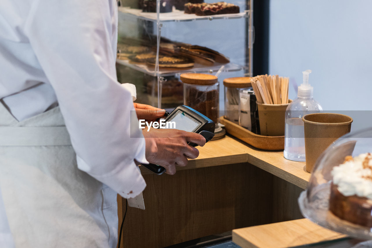 Side view of crop anonymous seller using pos terminal for payment while standing at counter in bakehouse with vegan desserts