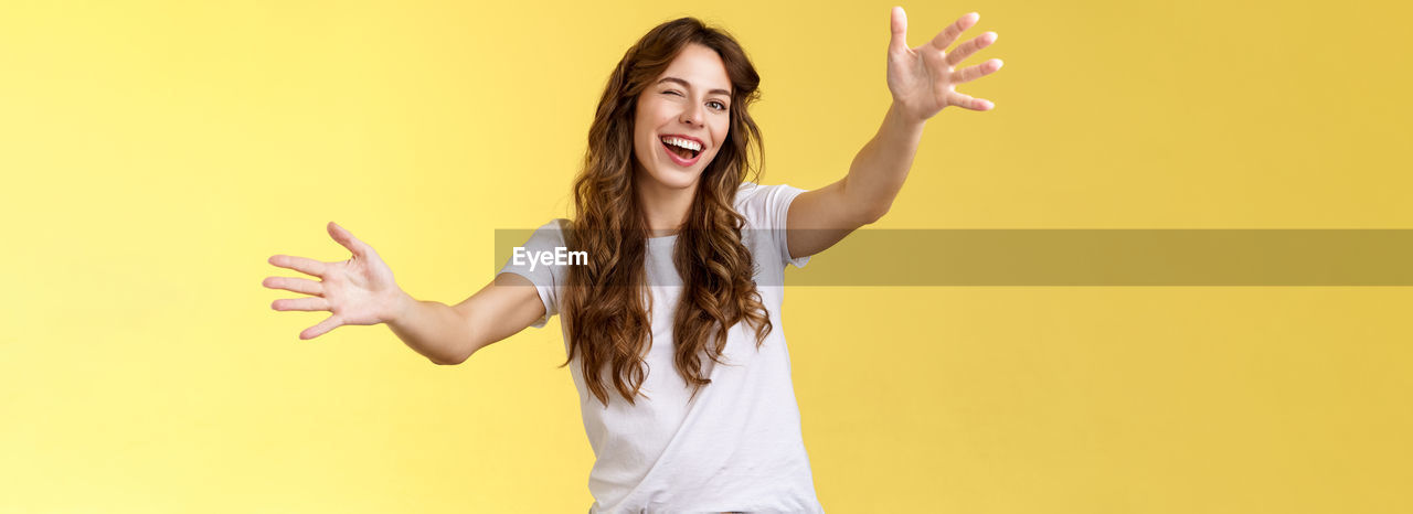 happiness, young adult, one person, women, smiling, studio shot, emotion, adult, yellow, long hair, colored background, arm, cheerful, positive emotion, hairstyle, fun, portrait, arms raised, limb, indoors, human limb, gesturing, sign language, waist up, joy, enjoyment, yellow background, brown hair, casual clothing, vitality, copy space, excitement, hand raised, hand sign, standing, carefree, finger, teeth, laughing, looking at camera, person, hand, smile, female, fashion, music, lifestyles, arms outstretched, arts culture and entertainment, shouting
