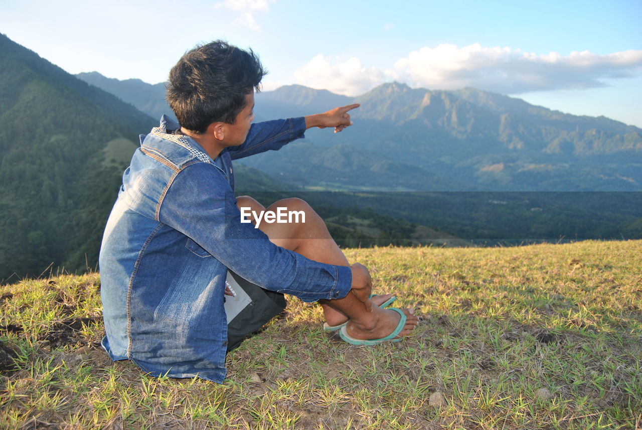 Young man pointing at mountain while sitting on cliff