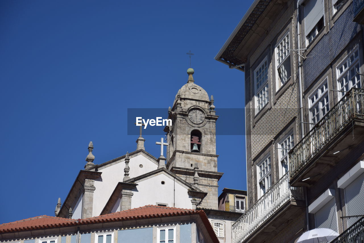 LOW ANGLE VIEW OF CLOCK TOWER AMIDST BUILDINGS AGAINST CLEAR BLUE SKY