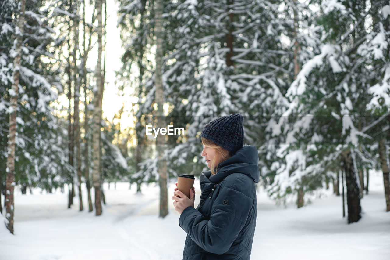 A nice middle-aged blonde woman walks through the winter forest with a cup of hot drink. 