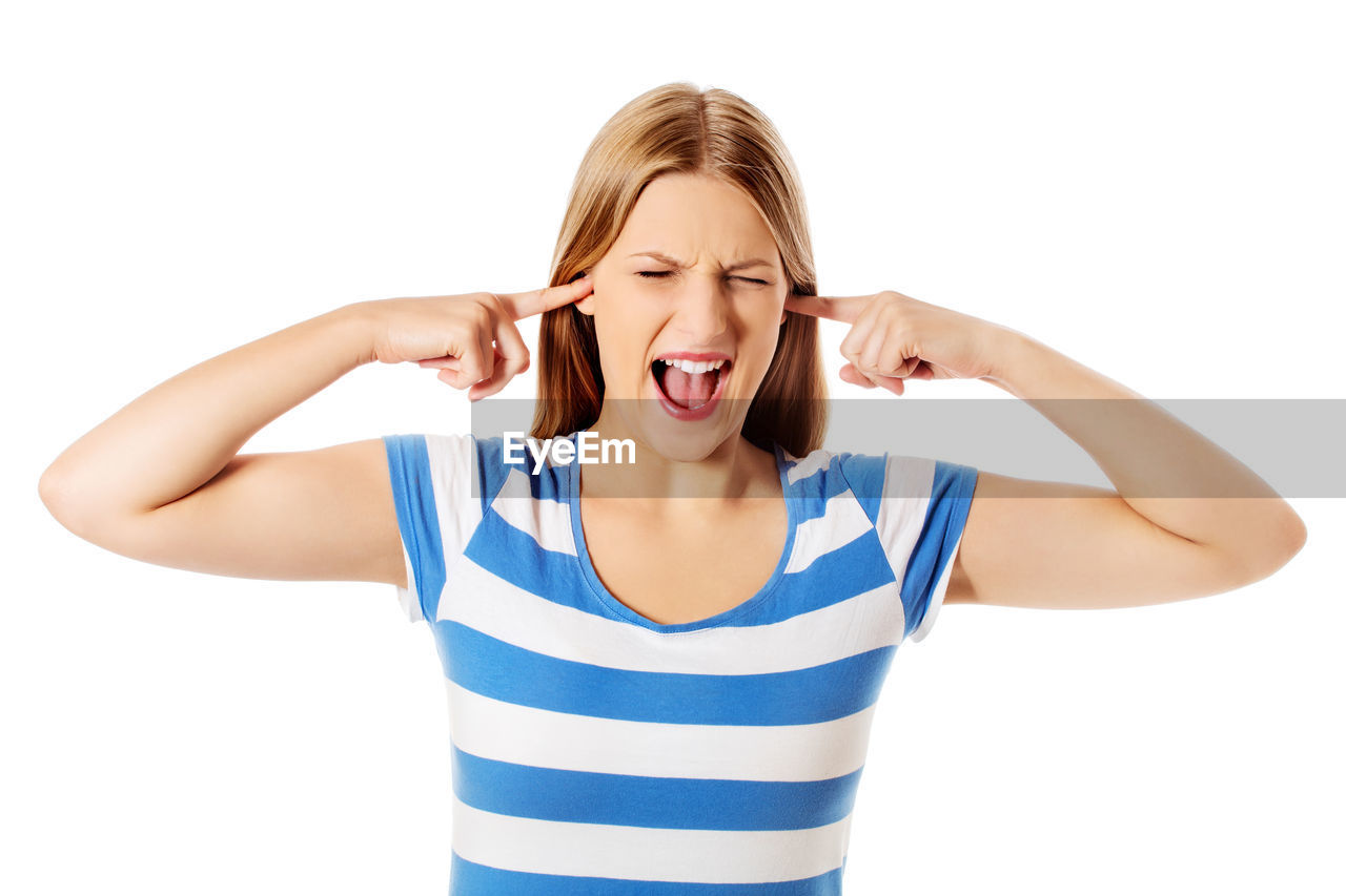 Young woman screaming while putting fingers in ears against white background