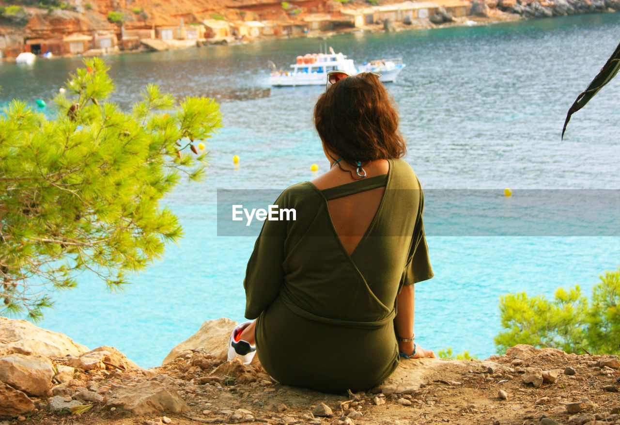 REAR VIEW OF WOMAN SITTING ON ROCK LOOKING AT SEA