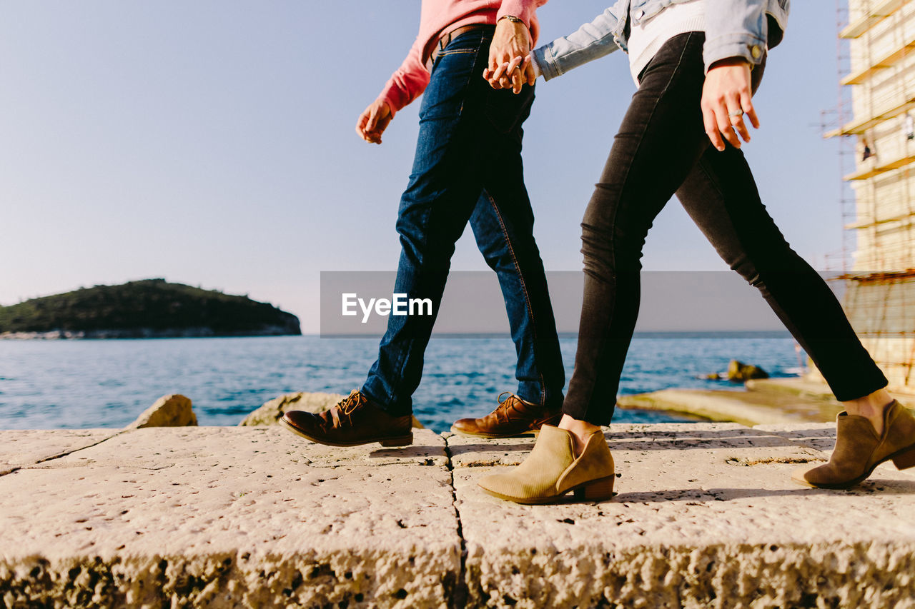 Low section of couple walking at beach against clear sky