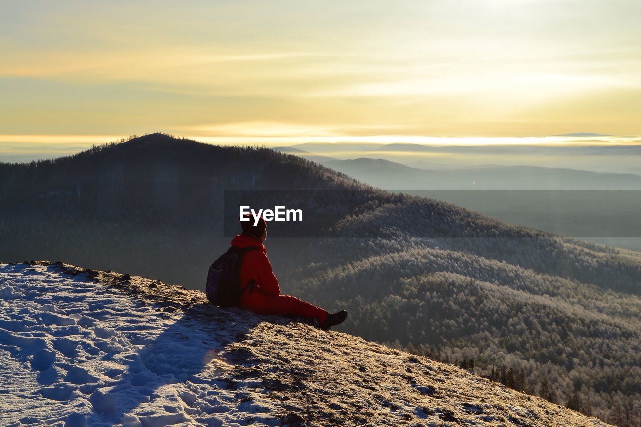 REAR VIEW OF MAN SITTING ON SNOWCAPPED MOUNTAIN DURING SUNSET