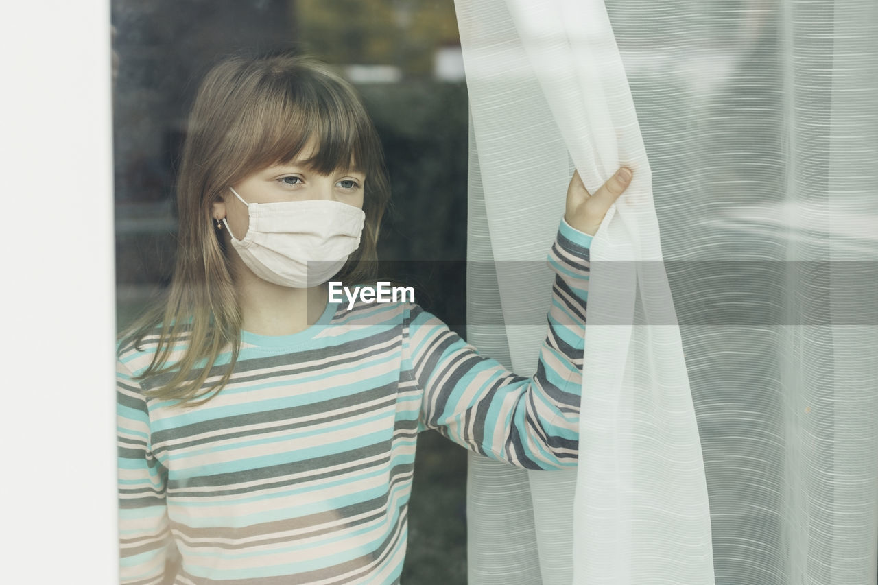 A school-age girl in a protective medical mask looks out the window. 