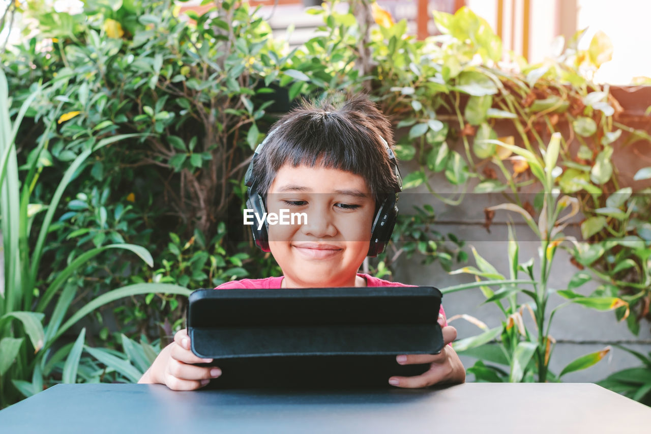 Asian cute boy wearing wireless headphones and smiling happily while playing games on the tablet.