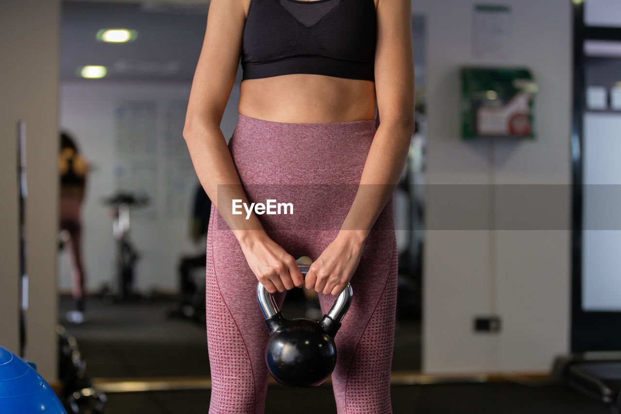 exercising, adult, gym, sports, lifestyles, sports clothing, sports training, indoors, women, wellbeing, clothing, one person, limb, standing, strength, sport venue, vitality, room, health club, exercise equipment, young adult, weight, arm, muscular build, focus on foreground, physical fitness, front view, athlete, human leg, fashion, midsection, determination, body conscious, weight training, female, sports bra, holding, self improvement, three quarter length, crop top