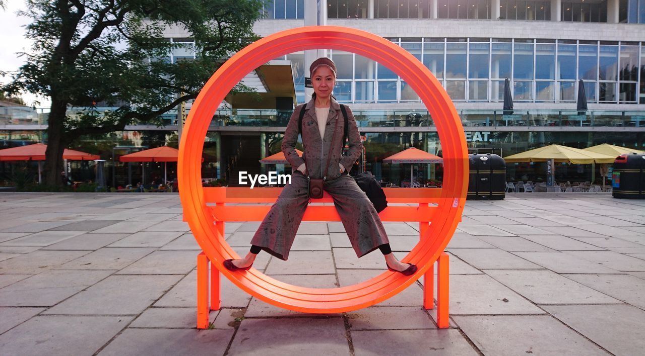 Full length portrait of woman sitting on orange structure against building in city
