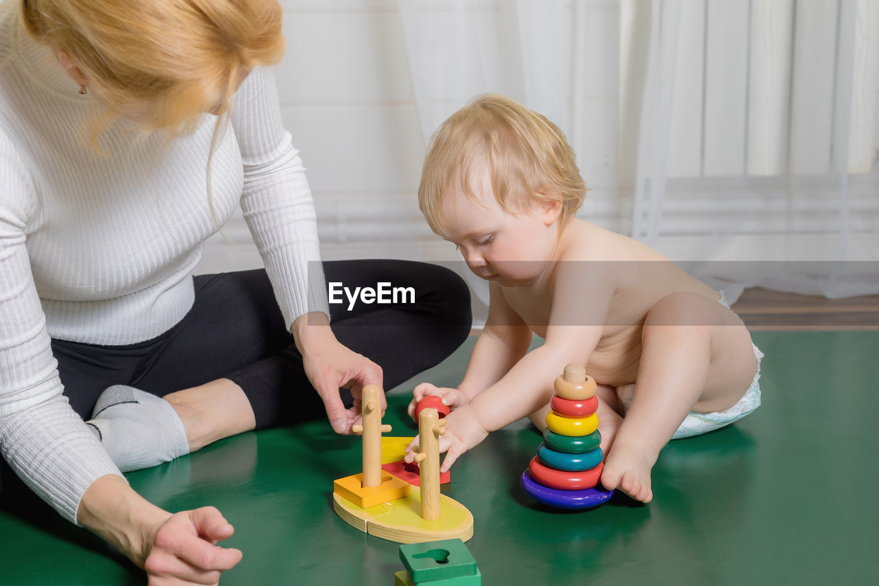 The kid sits on the rug, plays with his mother, collects a pyramid.