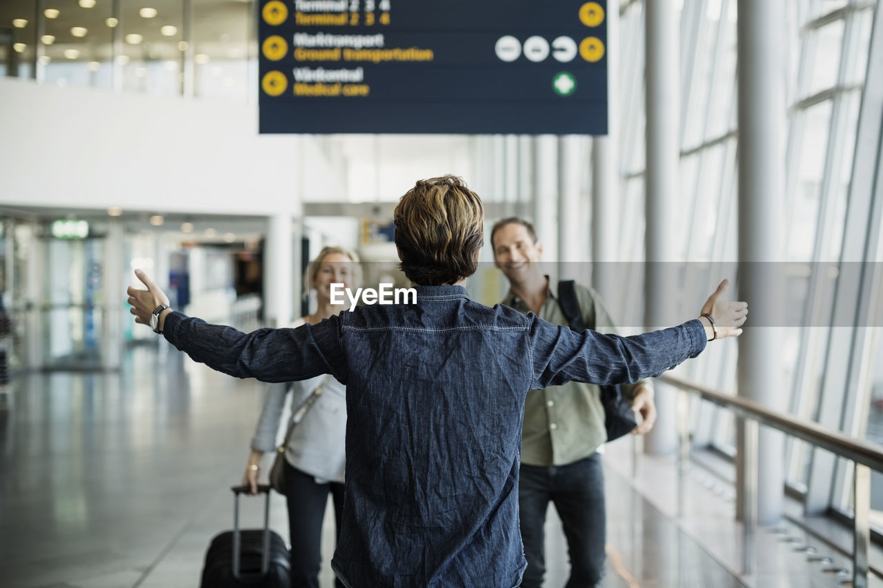 Rear view of businessman standing arms outstretched while greeting colleagues at airport