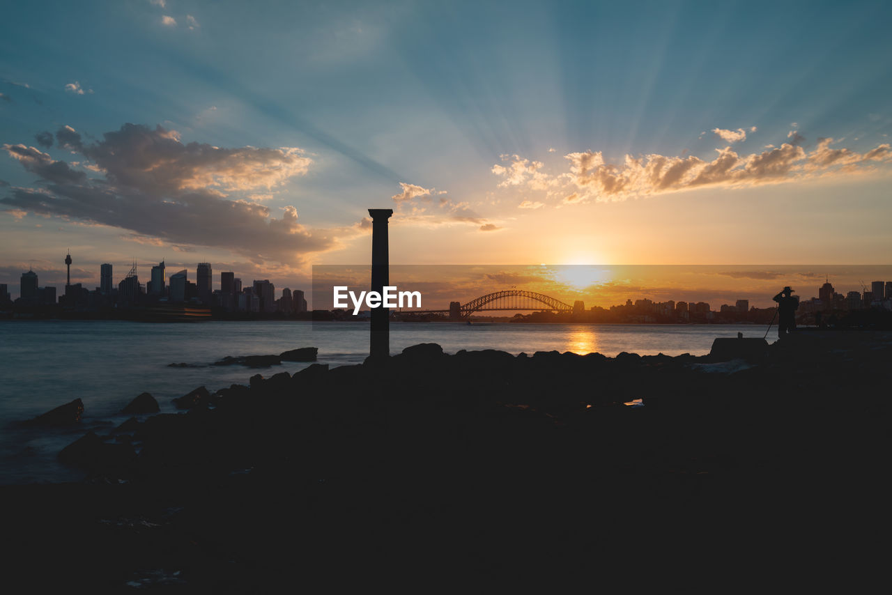 Silhouette buildings by sea against sky during sunset