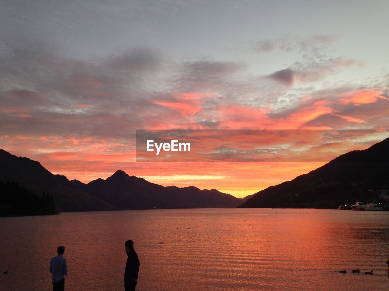 Men standing by lake against dramatic sky during sunset