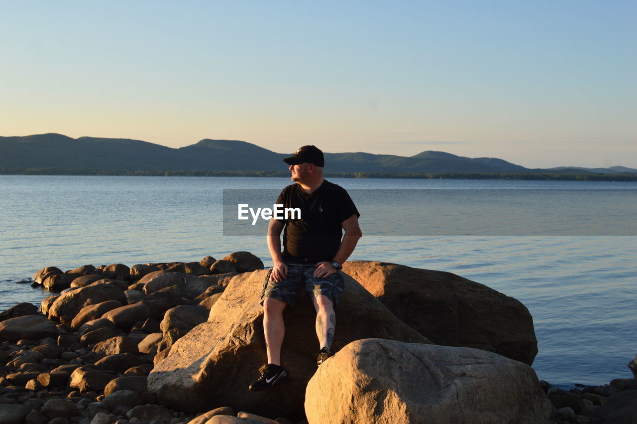 FULL LENGTH OF YOUNG MAN SITTING ON ROCK AT BEACH