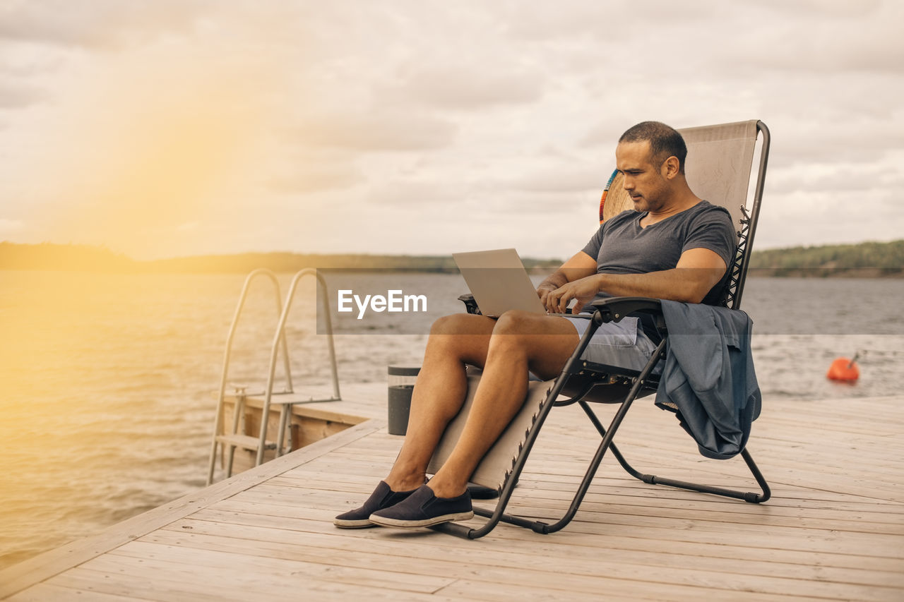 Full length of mature man using laptop while sitting on deck chair at jetty