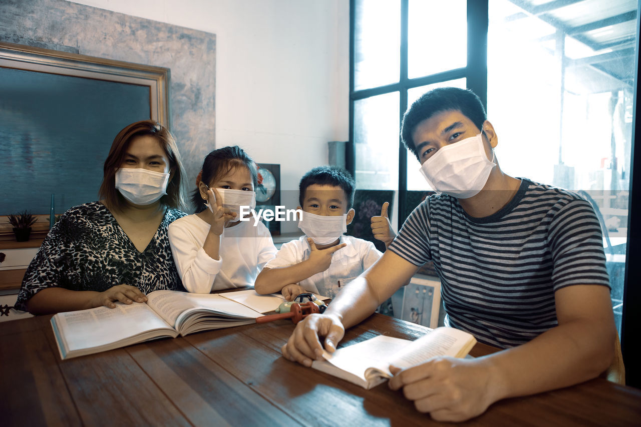 Portrait of cheerful family wearing mask sitting by table with book at home