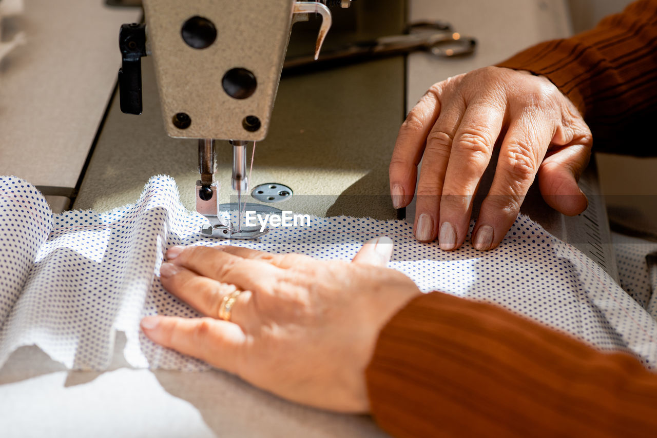 Cropped unrecognizable senior seamstress sitting at sewing machine and stitching piece of cloth while working in workshop