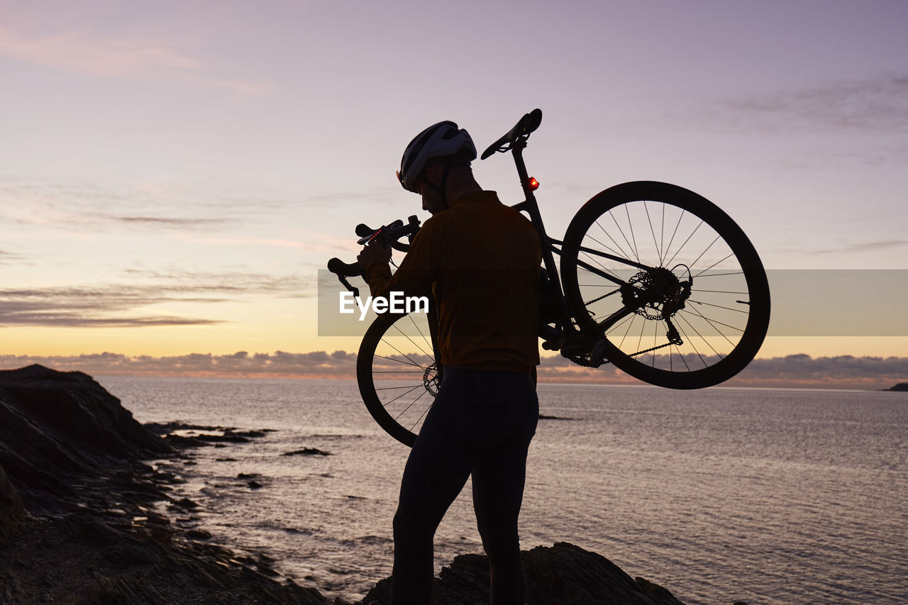 Cyclist carrying bicycle in front of sea at sunrise