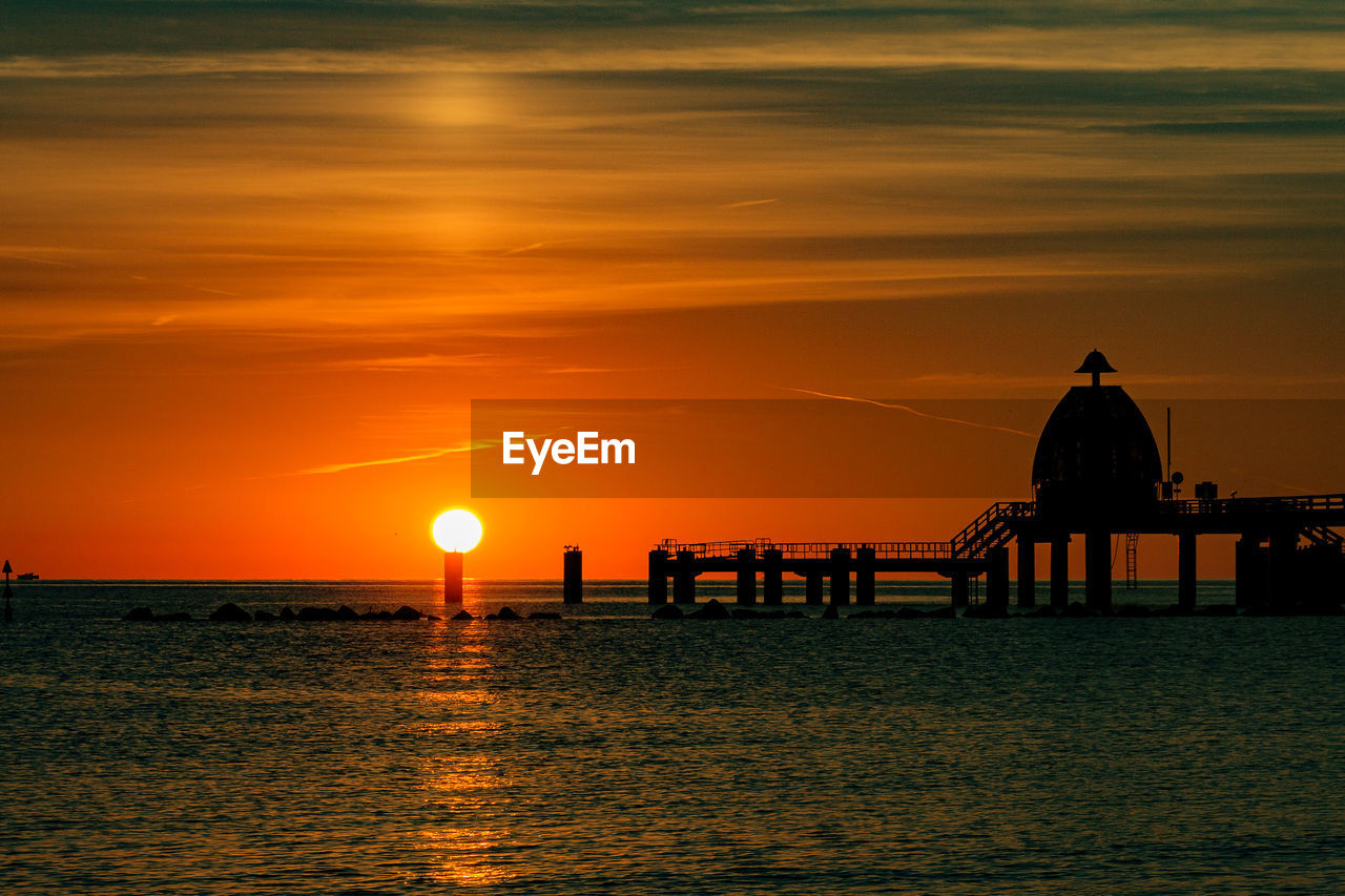 Silhouette built structure by sea against orange sky